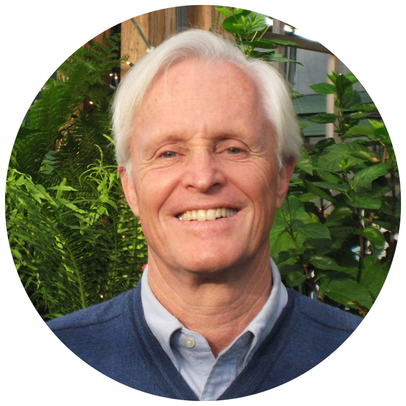 A white man with white hair, smiling at the camera. There are plants in the background.
