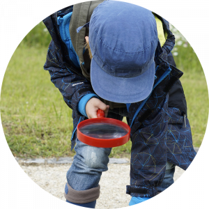 kid with blue hat outside bent over looking at the ground through magnifying glass
