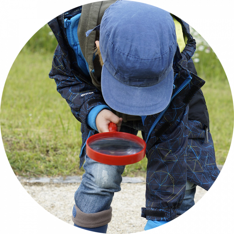 kid with blue hat outside bent over looking at the ground through magnifying glass