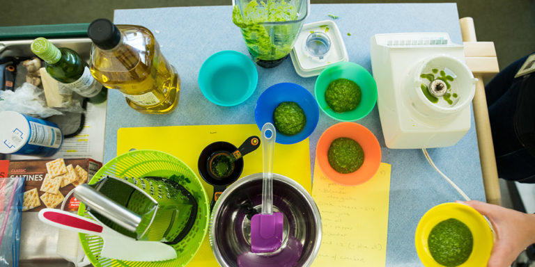 Cooking with students. Brightly colored bowls of pesto, plus a blender, cheese grater, cutting boards, knife, and crackers.