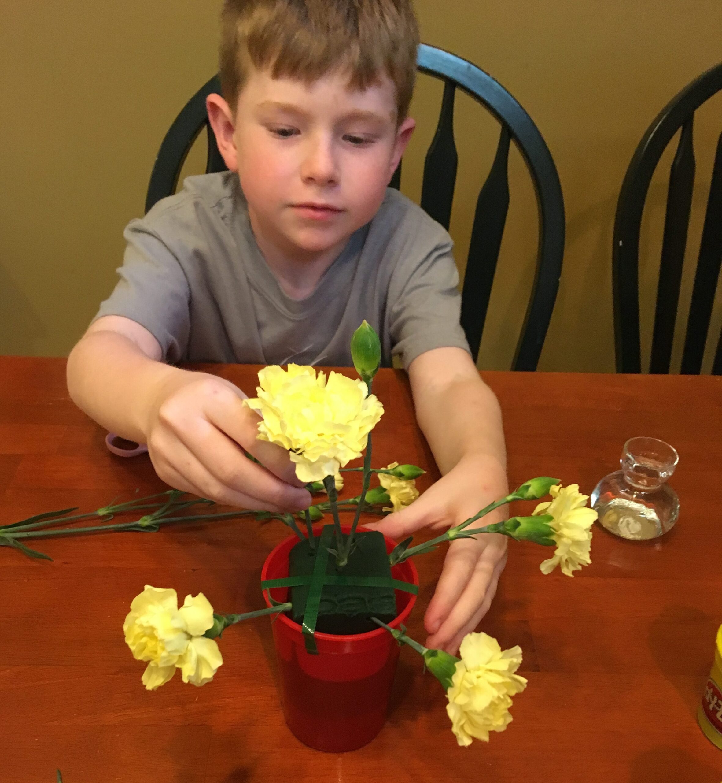 A boy pushing yellow carnation stems into floral foam in a red container