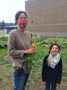 An adult in a face covering holds carrots standing next to a child with a carrot in their mouth