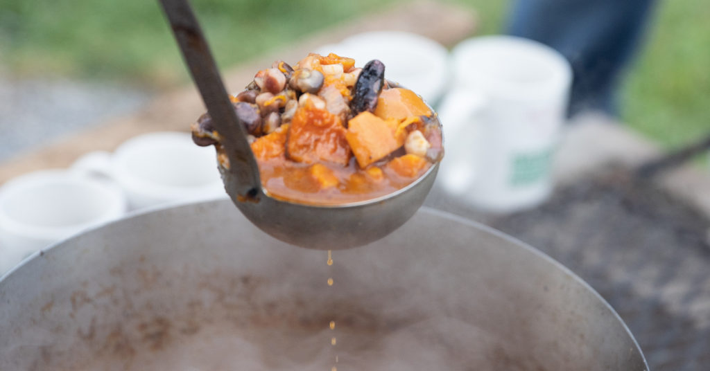 A soup ladle full of beans, squash, and other delicious food is about to be served into a mug with the Shelburne Farms logo