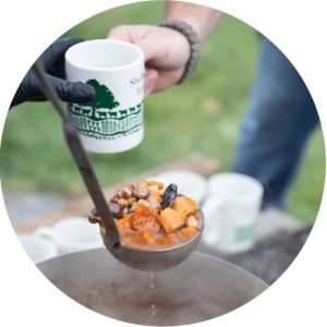A soup ladle full of beans, squash, and other delicious food is about to be served into a mug with the Shelburne Farms logo