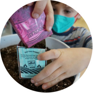 Child in a turquoise mask is holding two milk cartons over a bucket of soil