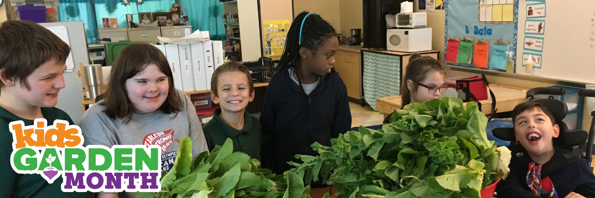 Kids Garden Month header A group of kids in a classroom looking happy near a table full of garden greens.