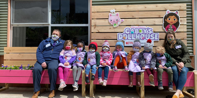 Children and adults sitting on a custom garden bench with the Gabby's Dollhouse logo and characters