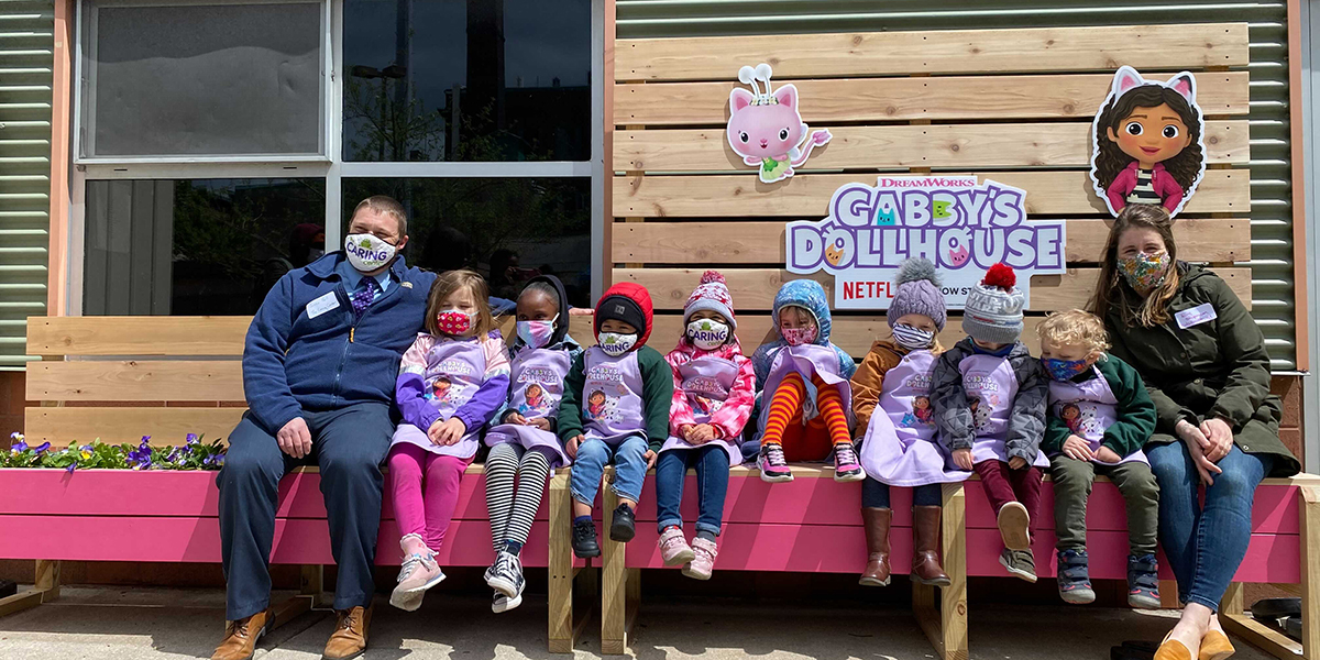 fairy tail blog header Children and adults sitting on a custom garden bench with the Gabby's Dollhouse logo and characters