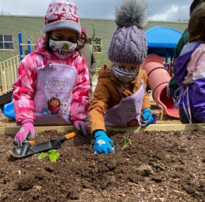 Kids in colorful hats, coats, face coverings, and garden gloves are planting in a raised bed.
