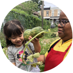 An adult and child harvesting a winter squash together. The child is smiling at the squash.