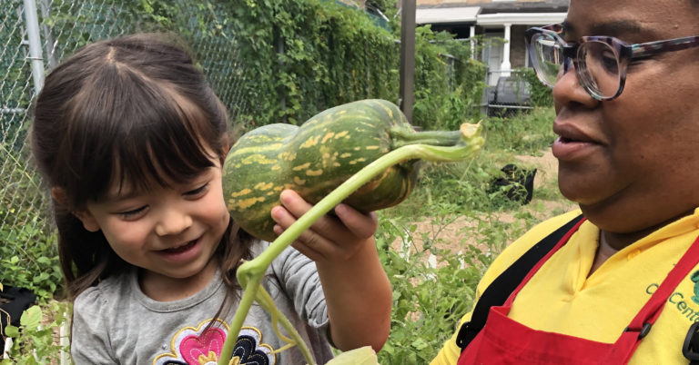 An adult and child harvesting a winter squash together. The child is smiling at the squash.
