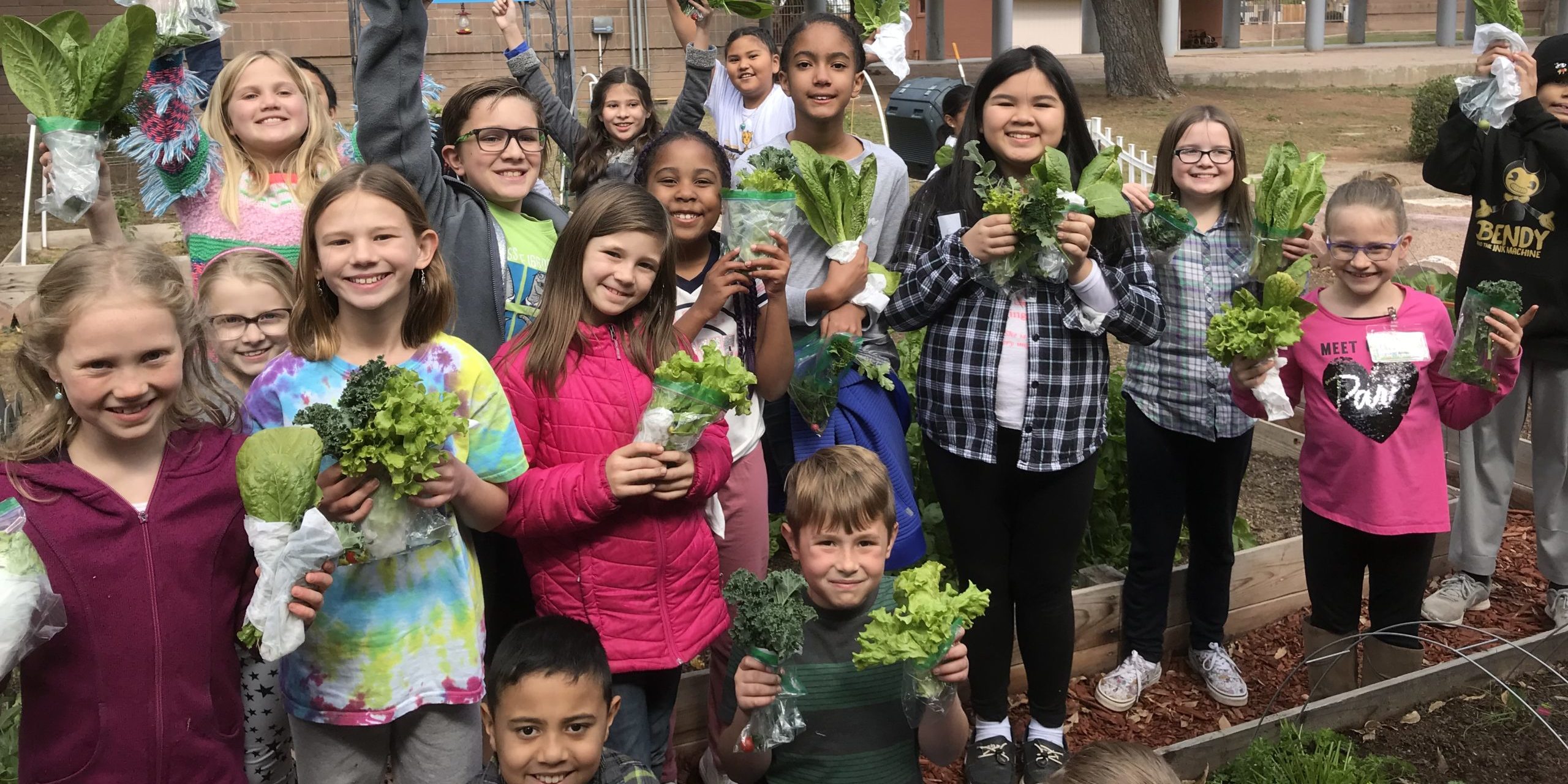 DF8B4DAE-2A92-47F1-B2AB-0C583D680BDF A diverse group of children smile and hold garden produce. They are outside, and a brick building is in the background.