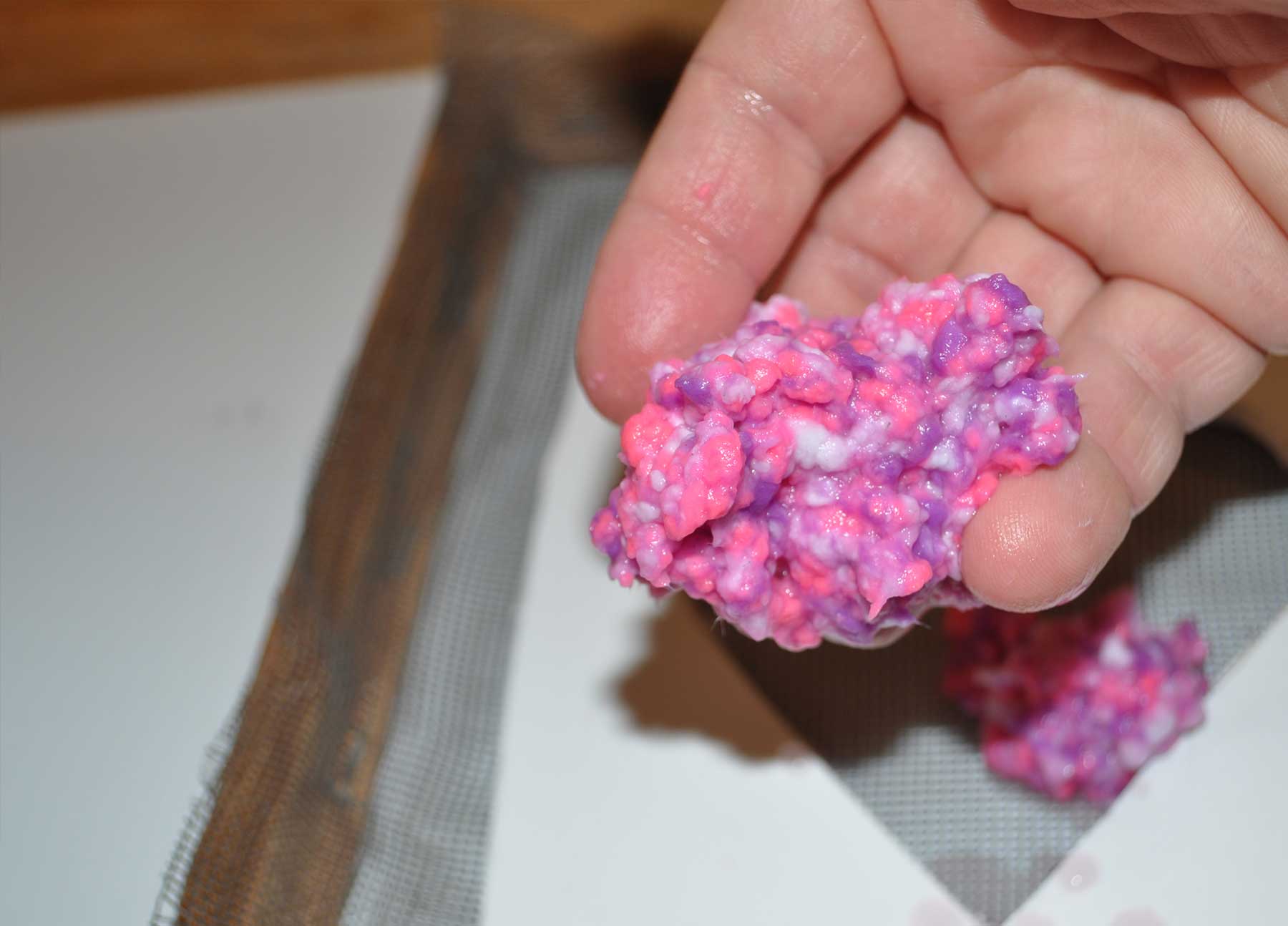 A hand holding a clump of mixed pink and purple paper pulp over the frame