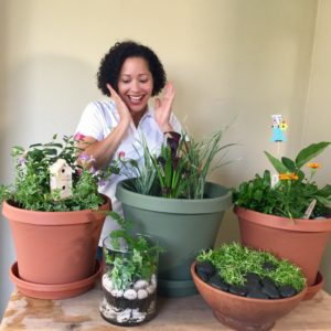 Perla, a woman with short dark curly hair, is holding her hands up to her face in surprise. In front of her are five planters with plants and fun whimsical elements such as tiny birdhouses and colorful stakes and flowers.