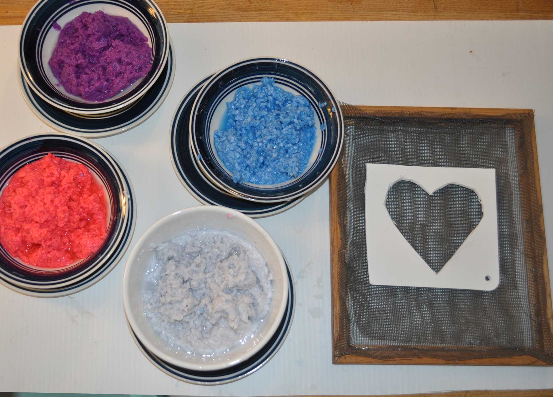 Bowls of red, blue, purple and white paper pulp next to the screen frame with a heart stencil