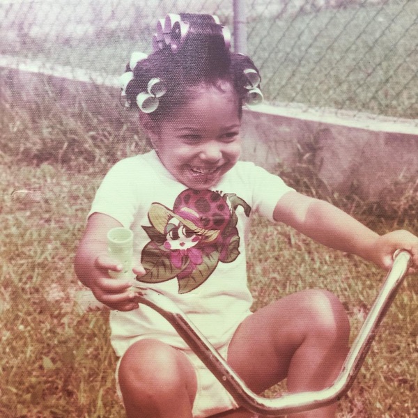 A toddler with curlers in her hair riding a tricycle. She's outside, and grass and a fence are visible in the background. The photo is old and has faded tones.