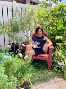 A woman with dark brown hair sits in a pink chair in a garden. She's smiling and a book is open on her lap. Behind her are pink flamingo decorations, small trees, and lush green plants.