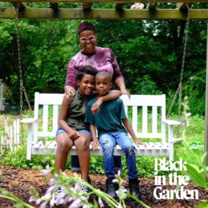 A Black family of an adult and two children hugging on a white bench in a garden. The words Black in the Garden are in the bottom right corner.