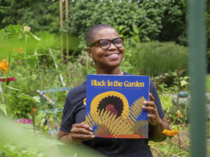 A Black person in a garden is holding a blue book titled Black in the Garden