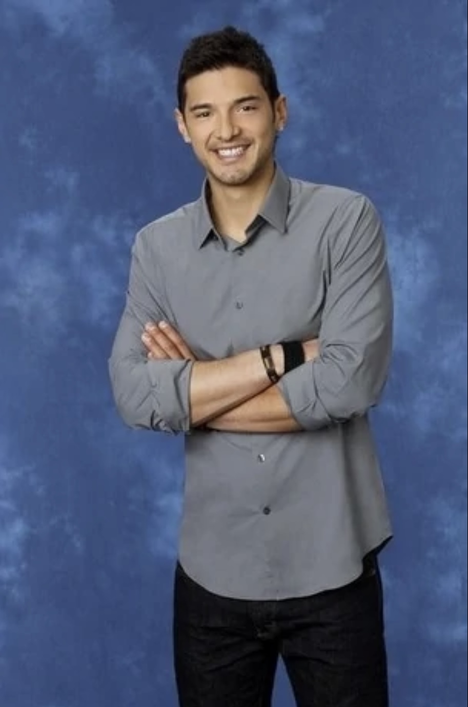 A 3/4 length shot of Alex, a man standing with his arms crossed in front of a blue background