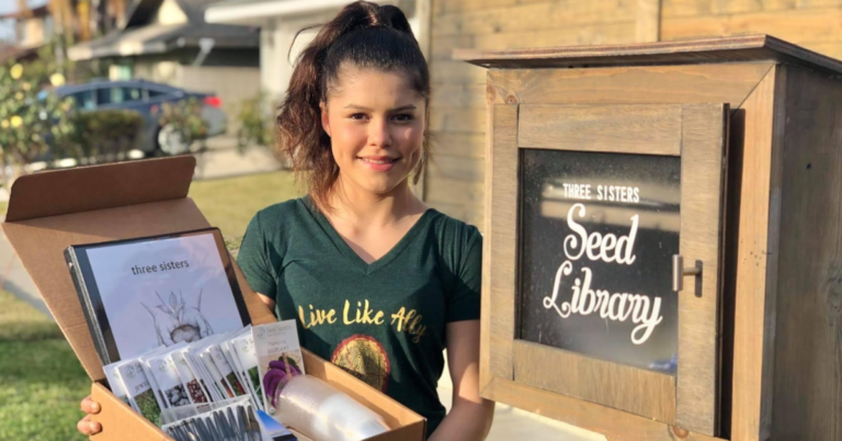 A teen holds a box of seeds in front of an outdoor cupboard that says Seed Library