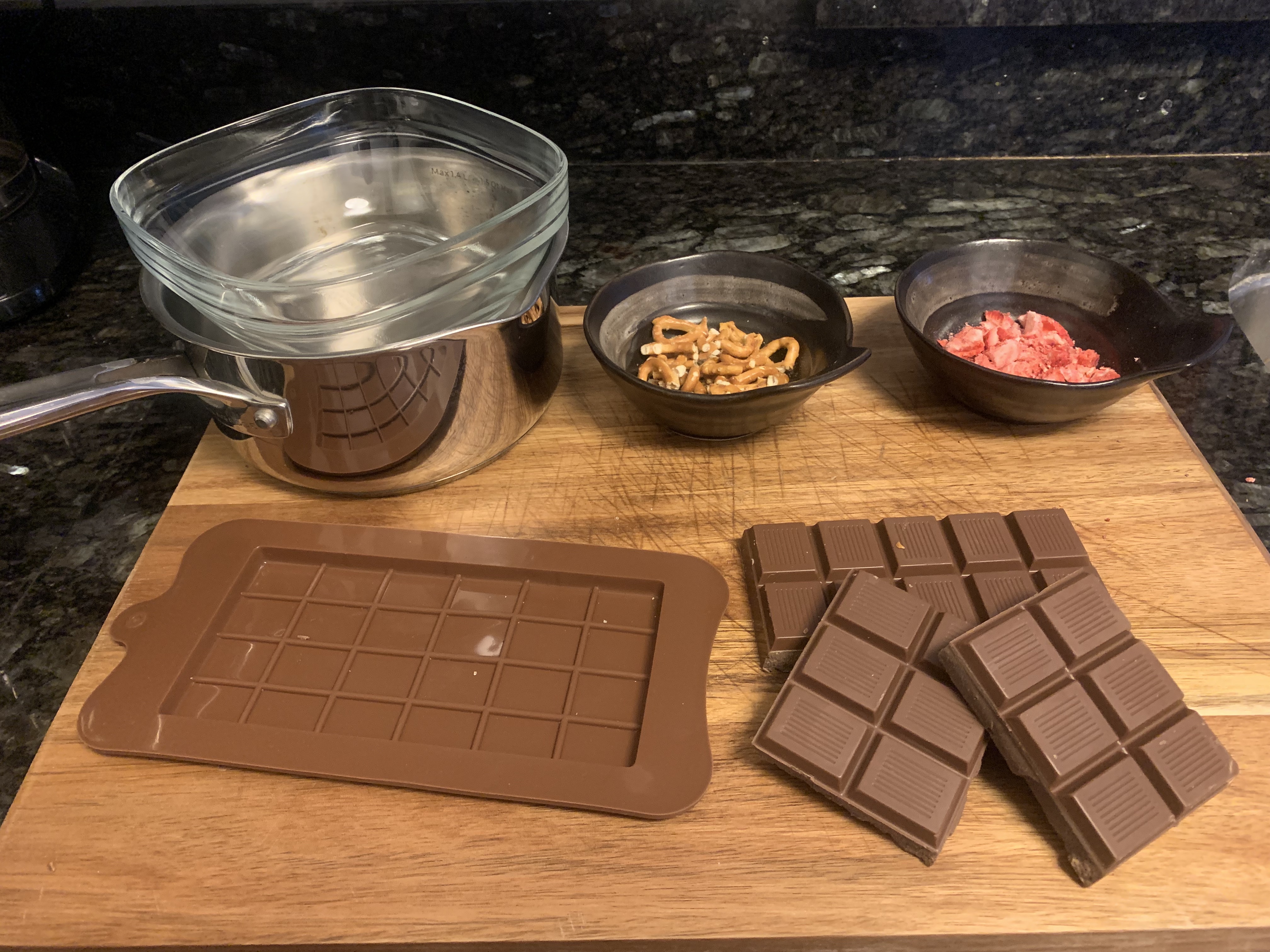 A wooden board with chocolate, a double boiler, and bowls of chopped ingredients