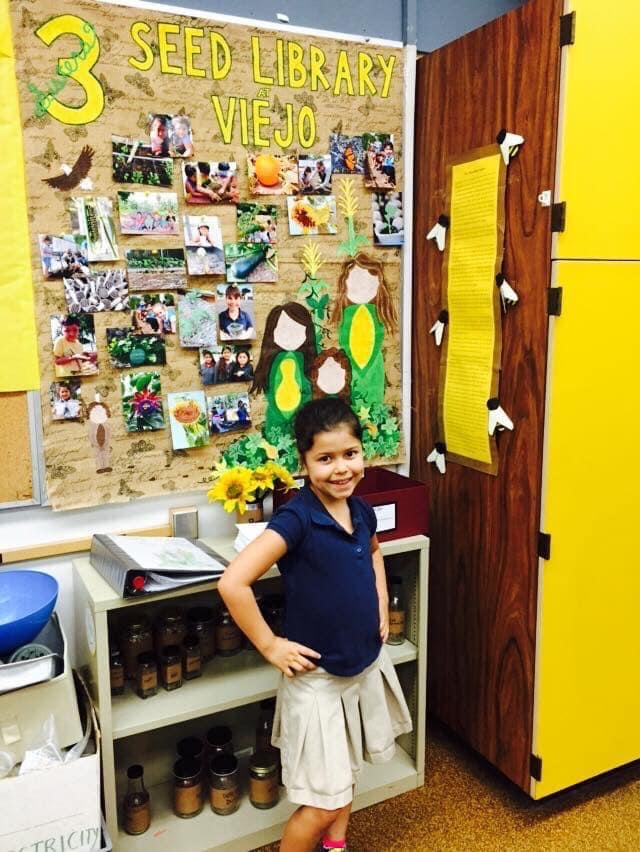 A child in a school uniform standing in front of a seed library on a bulletin board.