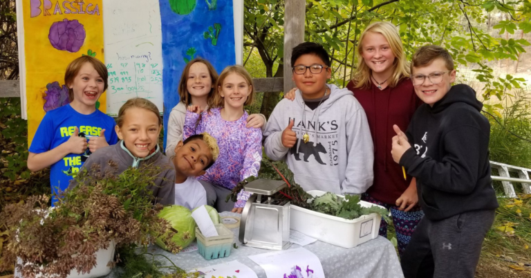 Seven kids of different ages standing in front of a table holding fresh produce