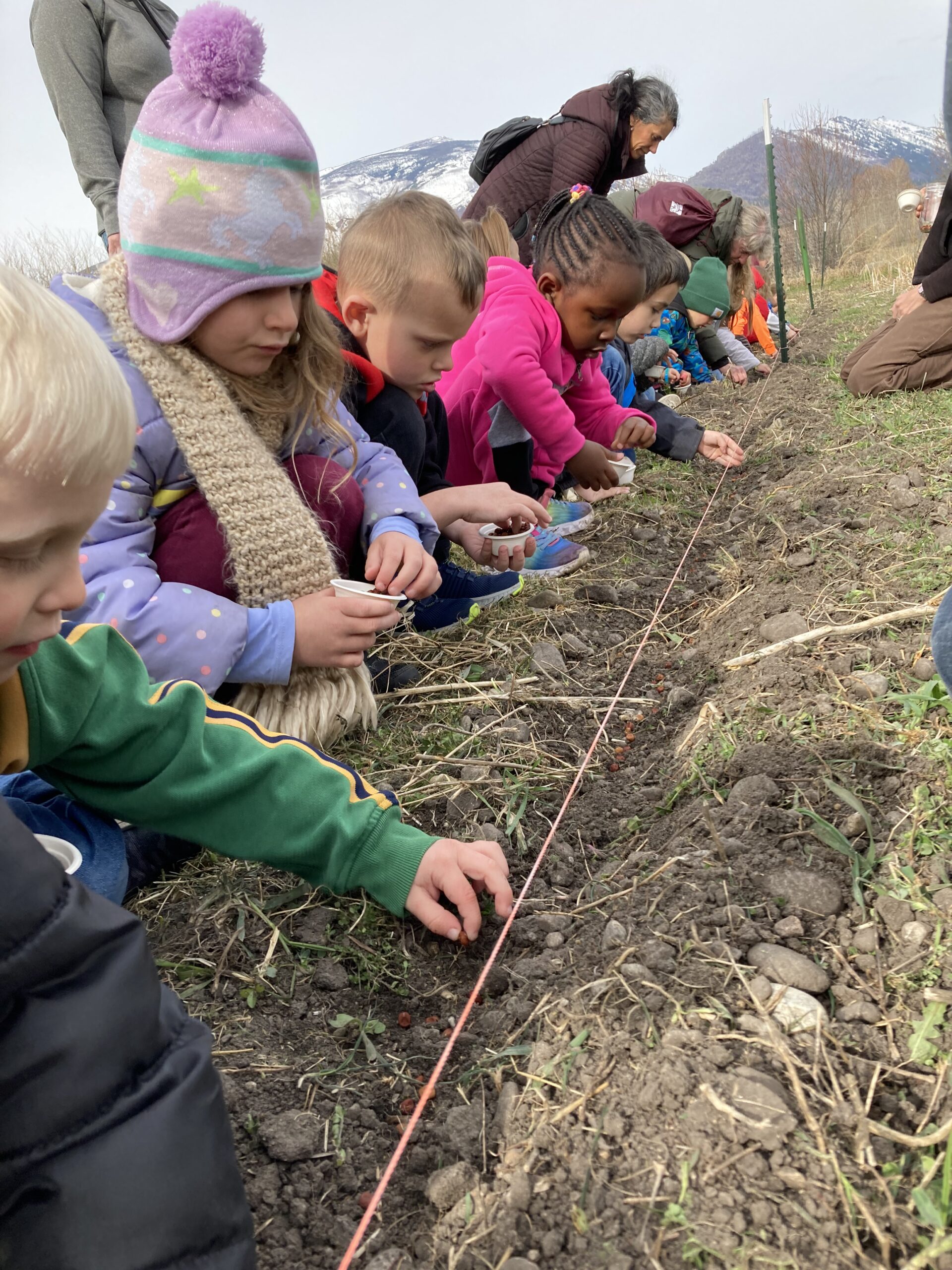 Kids squatting together in a line, in front of a narrow trench in which they are placing seeds
