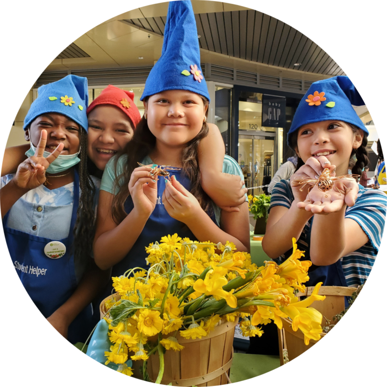 Children wearing gnome hats and smiling at the camera. A few are holding small items and one is giving the peace sign. A bouquet of yellow daffodils is in the foreground.