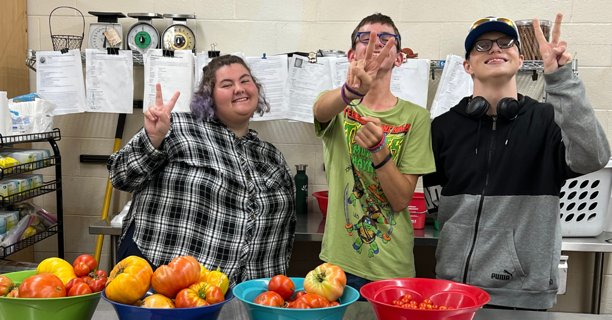 Three teenagers giving the peace sign while standing behind a table covered with colorful tomatoes.