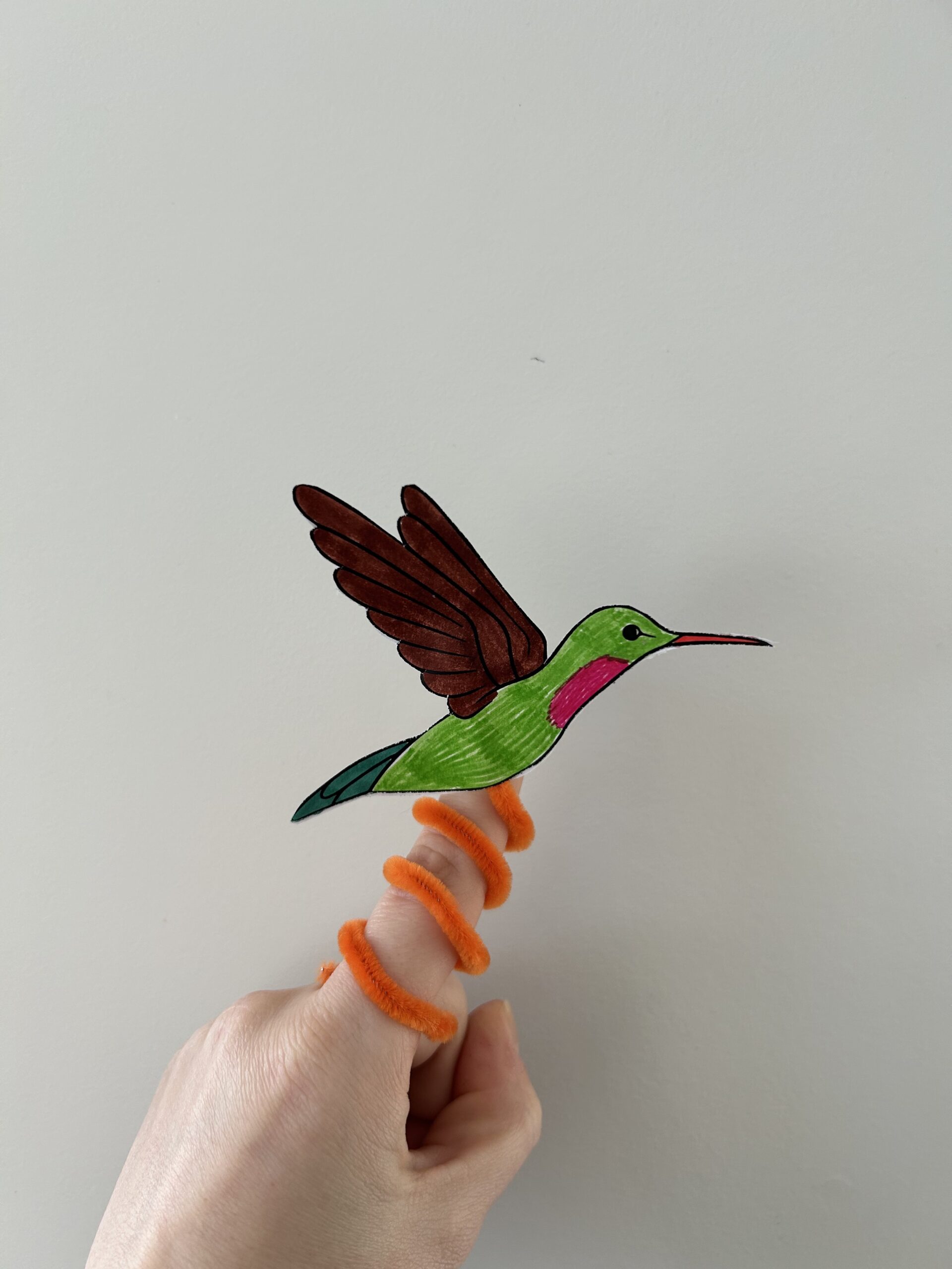 A colorful paper hummingbird attached to an orange pipecleaner, curled around a finger.