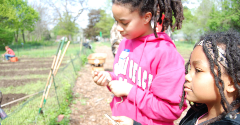 Two kids outside in a garden. ONe is wearing a pink sweatshirt and twists in their hair, the other has a black sweatshirt and twists in their hair.