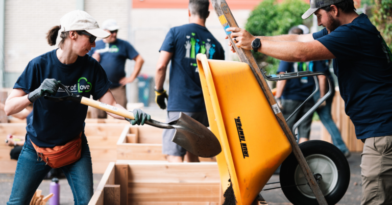A person in a blue t-shirt scoops soil out of a yellow wheelbarrow held by another person in a blue tshirt.