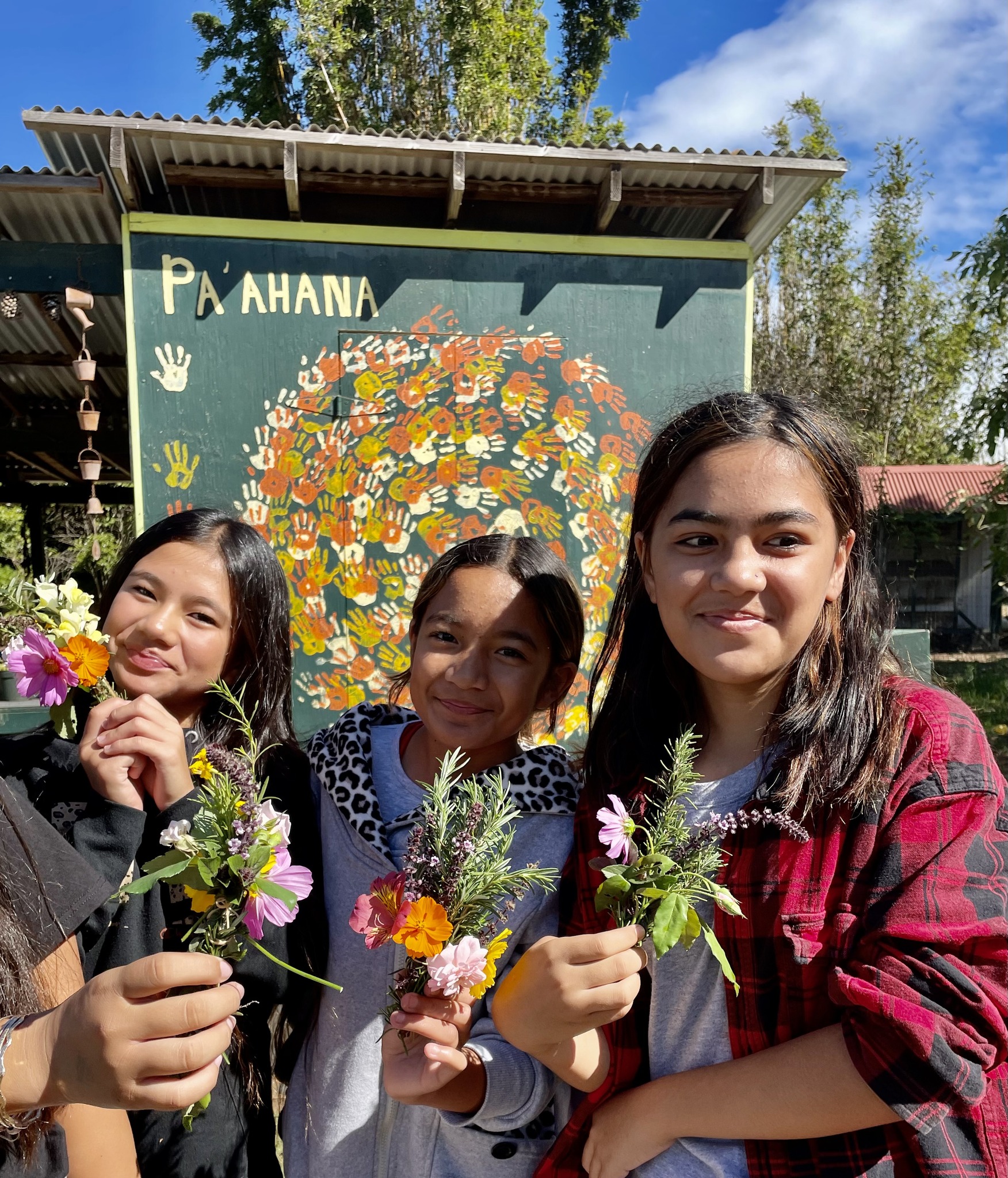 Three kids holding flowers outside in front of a sign.