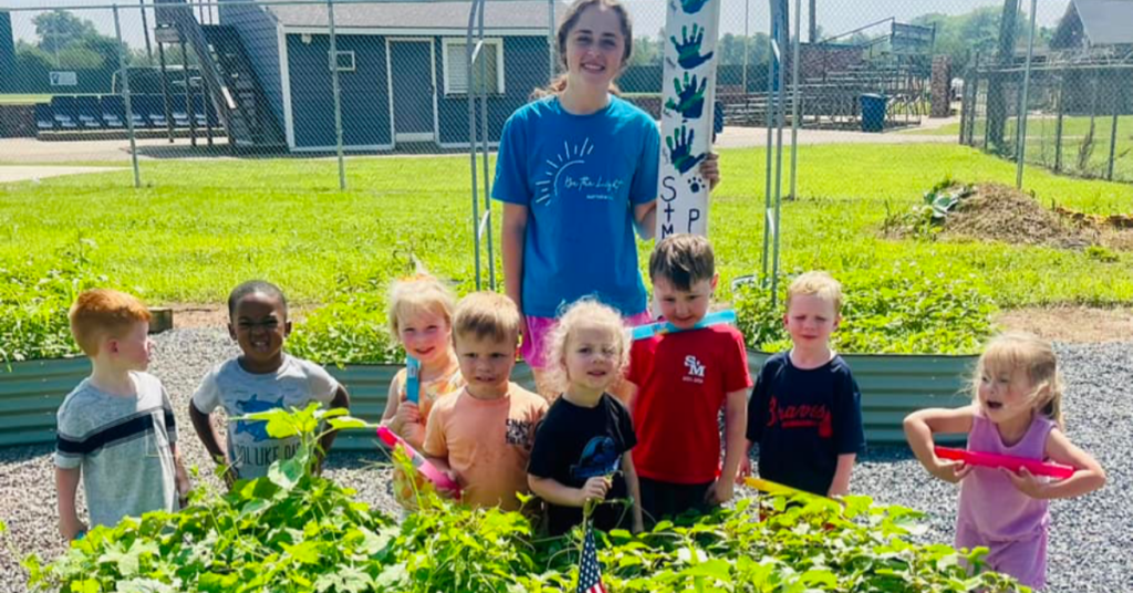 An adult stands behind eight children standing at a raised bed.