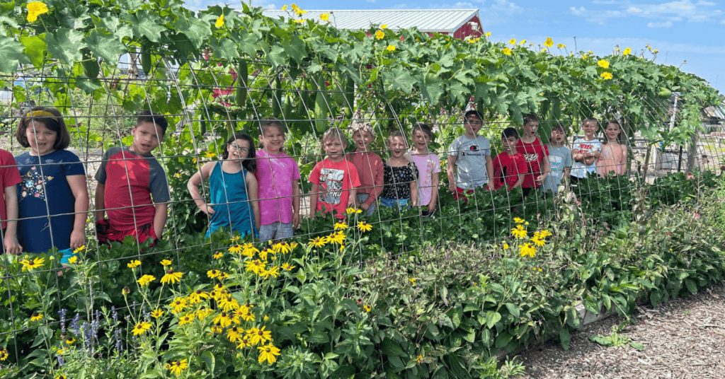 A long row of kids standing in a garden tunnel that has greenery and flowers growing on it.
