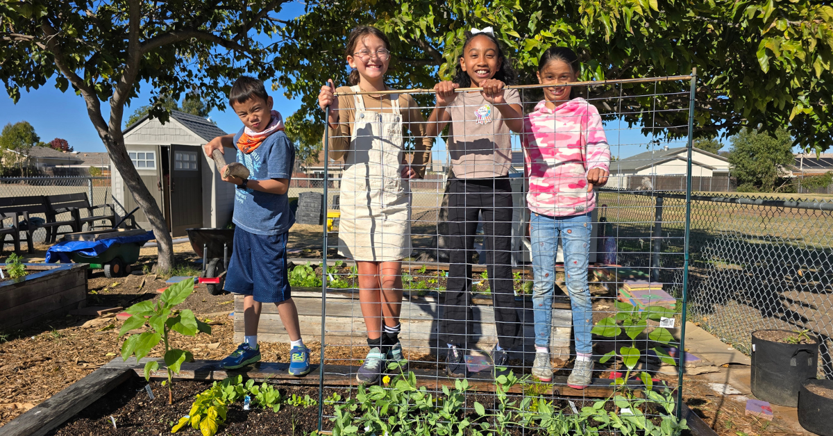 Four kids in a garden, three standing behind a trellis and one holding a log.